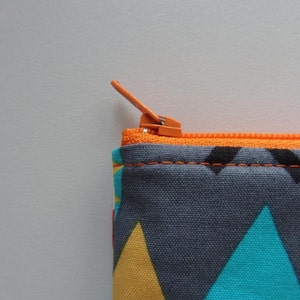 Può includere: Primo piano di una cerniera su una pochette in tessuto grigio con un motivo geometrico in giallo, blu e rosso. La cerniera è arancione.