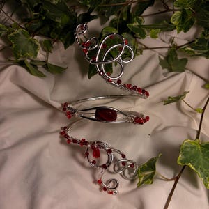 Può includere: Un bracciale da braccio in filo metallico color argento con un design a spirale decorato con piccole perline rosse e una singola pietra ovale rossa, liscia e più grande, incastonata al centro. Il filo è intrecciato in volute decorative.