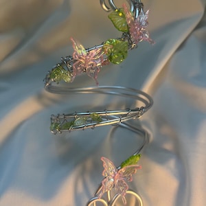 May include: A silver-toned arm cuff with a spiral design. The cuff is adorned with pink and clear butterfly accents and green leaf-shaped embellishments. The cuff wraps around the arm and features decorative swirls at the top and bottom.