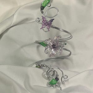 May include: A silver-coloured metal arm cuff with a spiral design. It features a purple butterfly, a translucent purple flower, and green leaves. The cuff is adorned with small, dark purple beads.