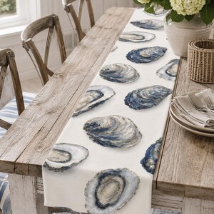 May include: A white table runner featuring a repeating pattern of oyster shells in shades of blue, gray, and cream. The shells are depicted with detailed textures and varied shapes, arranged across the length of the fabric runner.