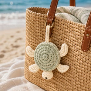 May include: A beige woven tote bag with brown leather handles and a crochet turtle keychain attached to one handle. The turtle features a sage green circular shell, cream-colored flippers, and a small head with a single black eye detail.