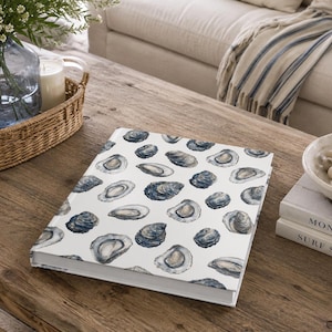 May include: A hardcover book featuring a repeating pattern of oyster shells on a white background. The shells are rendered in shades of dark blue, grey, and white, showcasing various angles and textures of the exterior and interior of the shells.