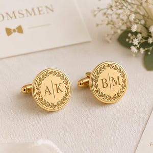 May include: Gold-colored cufflinks with engraved initials inside a laurel wreath design. The image includes a card that says "Groomsmen" and a tag that says "Thank you for standing by my side". A white rose and ribbon are also in the image.