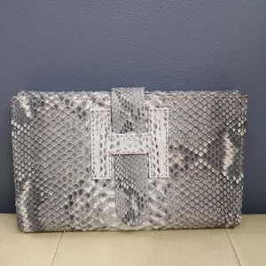 Puede incluir: Un bolso de mano rectangular con un estampado de piel de serpiente en tonos grises y blancos. Cuenta con una correa de cuero blanco y un cierre de cuero blanco con forma de letra "H".