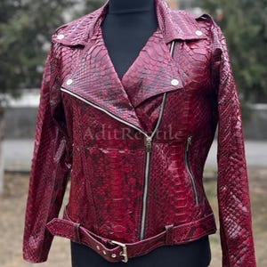 May include: A burgundy leather jacket with a snakeskin pattern. The jacket features a notched collar, an asymmetrical zipper closure, and a belt at the waist. The sleeves are long, and the jacket is displayed on a black mannequin.