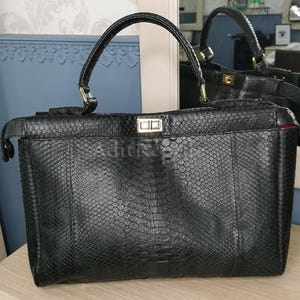 May include: Black leather handbag with a textured snakeskin pattern. The bag has a rectangular shape with a top handle and a gold-colored clasp. The interior is lined with red fabric. The bag is reflected in a mirror.
