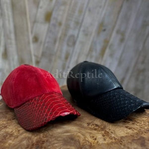 Puede incluir: Se muestran dos gorras de béisbol, una roja y otra negra. La gorra roja tiene una corona de ante y una visera con textura de piel de serpiente. La gorra negra es de cuero con una visera con textura de piel de serpiente.