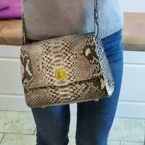Puede incluir: Bolso bandolera rectangular con estampado de piel de serpiente en tonos grises, negros y crema. El bolso presenta un cierre y herrajes dorados. La correa es del mismo material de piel de serpiente.