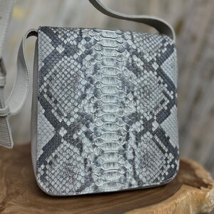 May include: A square, gray and white snakeskin-patterned crossbody bag with a matching gray strap. The bag is made of a textured material and has a flap closure. The bag is sitting on a wooden surface.