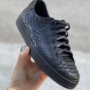 Pode incluir: Tênis pretos com um padrão texturizado em forma de escamas. Os sapatos têm cadarços pretos e sola preta. Os tênis parecem ser feitos de couro ou material semelhante e são de estilo baixo.