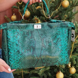 May include: A teal green handbag with a snakeskin pattern. The bag has a rectangular shape with a top handle and a silver clasp closure. The bag is held up in front of a Christmas tree with gold ornaments.