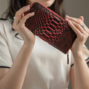 Puede incluir: Una cartera de cuero rojo y negro con un estampado de piel de serpiente. La cartera tiene cierre de cremallera y una borla. La cartera está sujeta por una persona.