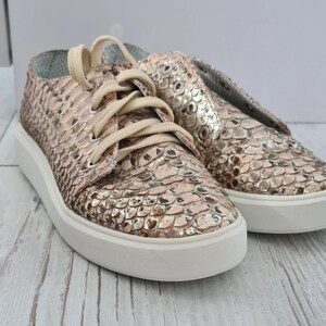 May include: Metallic rose gold sneakers with a snakeskin design. The shoes feature cream-coloured laces and a thick white sole. The sneakers are displayed on a light-coloured wooden surface.