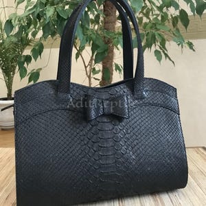 May include: Black tote bag with a bow detail. The bag has a textured, scaly pattern and two top handles. The text "AditRept" is visible on the front of the bag. The bag is sitting on a wooden surface.