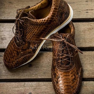 May include: A pair of brown leather sneakers with a snakeskin pattern. The shoes have white soles and brown laces. The sneakers are displayed on a wooden surface.