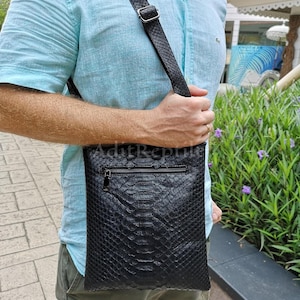 May include: Black leather crossbody bag with a textured snakeskin pattern. The bag has a zippered main compartment and a long, adjustable strap. The bag is being held by a person wearing a light blue shirt.