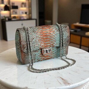 May include: A rectangular handbag with a snakeskin pattern in teal, orange, and brown tones. The bag features a silver chain strap and a silver clasp closure. The bag is sitting on a white marble table.