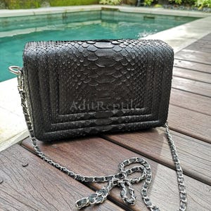 May include: A black, rectangular handbag with a silver chain strap. The bag is made of a textured material, possibly snakeskin, and features the text "Adit Reptile". The bag is sitting on a wooden surface.