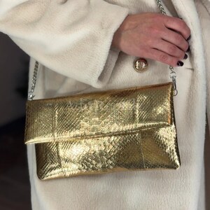 May include: A gold clutch with a chain strap. The clutch has a textured, snakeskin-like pattern and a fold-over flap. The chain is silver. The background is a cream-colored coat.