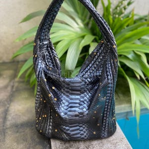 May include: Black snakeskin-textured handbag with a single shoulder strap. The bag features a rounded shape and is adorned with small, gold-colored studs. The background includes green foliage and a blue pool.