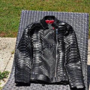 May include: A black motorcycle jacket featuring a textured, scale-like pattern across the entire exterior. The jacket includes a silver-toned asymmetrical front zip, a collared neckline, a buckled waist belt, and a vibrant red interior lining.