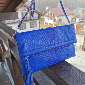 May include: A vibrant blue clutch with a snakeskin texture, featuring a silver chain strap. The bag has a zipper closure and a matching tassel. The clutch is displayed outdoors on a wooden railing.