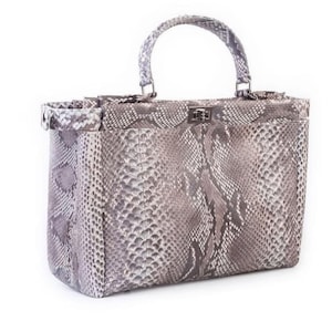 May include: A rectangular handbag with a top handle, featuring a snakeskin pattern in shades of gray and white. The bag has a silver-colored clasp and hardware. The handle is curved and attached to the top of the bag.