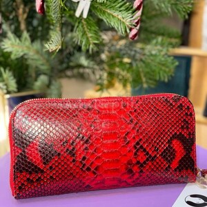 Puede incluir: Una cartera roja y negra con textura de piel de serpiente y cierre de cremallera. La cartera es rectangular y tiene una correa de muñeca. El fondo incluye un árbol de Navidad con decoraciones de bastones de caramelo.