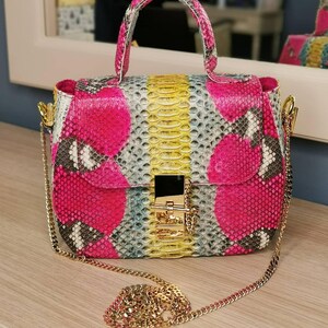 May include: A colorful handbag with a snakeskin pattern. The bag features a top handle and a gold chain strap. The main colors are pink, yellow, and gray. The bag has a gold clasp closure.