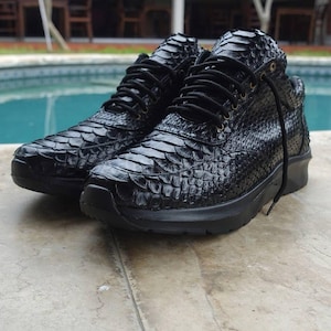 May include: A pair of black high-top sneakers with a textured, scaly pattern, resembling snake or reptile skin. The shoes have black laces and soles, creating a monochromatic look. The sneakers are placed on a light-colored surface.