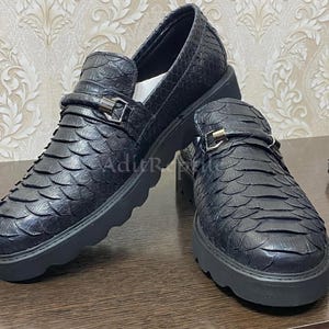 Può includere: Un paio di mocassini in pelle nera con un motivo strutturato a scaglie. Le scarpe hanno un dettaglio con fibbia e una suola spessa nera. I mocassini sono esposti su una superficie di legno.