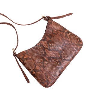 May include: A brown leather shoulder bag with a snakeskin pattern. The bag has a long, thin strap and a curved shape. The bag is made of a textured material and has a gold-colored clasp.