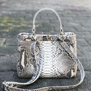 May include: A rectangular handbag with a snakeskin pattern in shades of gray and white. The bag has a top handle and a long, adjustable shoulder strap. A silver clasp secures the top flap.