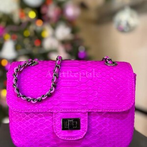 May include: A vibrant fuchsia handbag with a textured, reptile-like pattern. The bag features a silver chain strap and a rectangular clasp. The text "Art Reptile" is visible on the bag. The background is blurred with Christmas tree decorations.