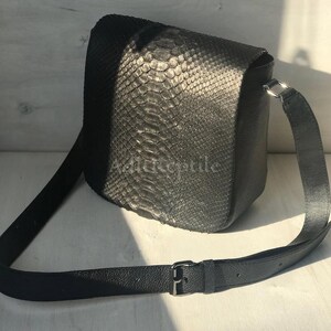 May include: Black crossbody bag with a textured snakeskin pattern. The bag has a long, adjustable strap and a silver-colored buckle. The bag is made of a dark, possibly leather material.