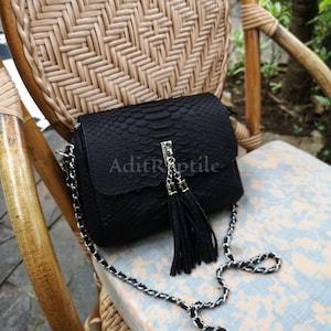 May include: Black crossbody bag with a textured snakeskin pattern. The bag features a silver chain strap and a decorative tassel. The text "AditRoptile" is printed on the front of the bag.
