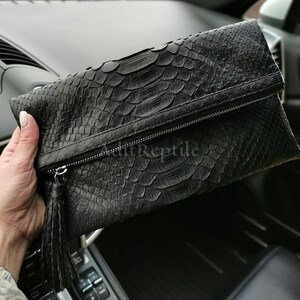 May include: Black clutch bag with a reptile-like texture. It features a zip closure and a tassel. The text "AuttReptile" is visible on the front.