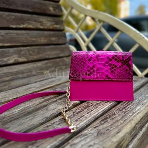 May include: A fuchsia-colored clutch with a snakeskin flap and a matching strap. The bag has a gold chain and is set on a wooden bench. The top flap is a vibrant pink snakeskin pattern, and the bottom is a solid fuchsia color.