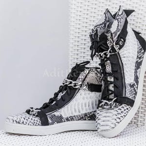 Könnte beinhalten: High-Top-Sneaker mit Schlangenmuster in Schwarz, Weiß und Grau. Die Sneaker haben schwarze Schnürsenkel, Reißverschlüsse und ein silbernes Kettendetail. Die Sohlen sind weiß.