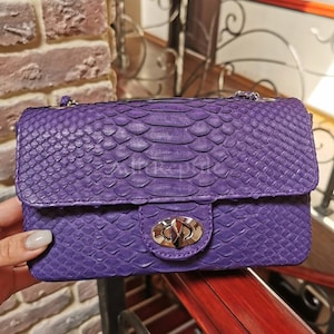 May include: A small, rectangular, purple handbag with a flap closure. The bag is made of a snakeskin-like material and features a silver-colored clasp. A silver chain strap is visible.