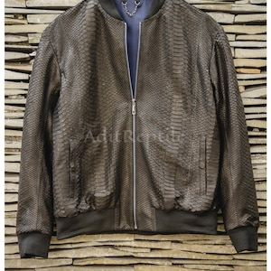 Peut inclure: Un blouson aviateur marron foncé avec un motif texturé en peau de serpent, une fermeture éclair centrale argentée, des poignets et un ourlet côtelés noirs, et une doublure intérieure bleue avec une chaîne argentée au col. Le texte AditReptile est visible.