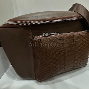 May include: A brown leather fanny pack with a textured snakeskin-like front pocket. The bag has a zipper closure and a brown strap. The text "AditReptile" is visible on the front.