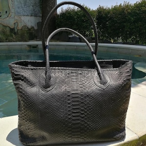 May include: A large, black tote bag with two handles. The bag is made of a textured material, possibly leather or a similar material, with a scale-like pattern. The handles are curved and attached to the top of the bag.