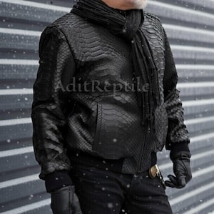 May include: Black leather jacket with a snakeskin texture, a black scarf, and black gloves. The jacket has a zipper closure and ribbed cuffs. The text "AditReptile" is visible on the jacket. The overall theme is fashion.