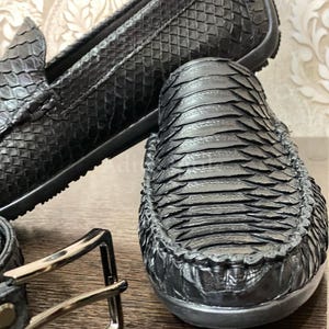 May include: Black leather loafers with a textured snakeskin pattern. The image also shows a matching black leather belt with a silver buckle. The shoes are displayed on a wooden surface.