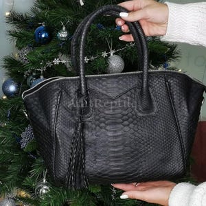 May include: A black handbag with a snakeskin pattern, featuring a top zipper, dual handles, and a decorative tassel. The bag is held in front of a Christmas tree with blue and silver ornaments. The text "Any Reptila" is visible.