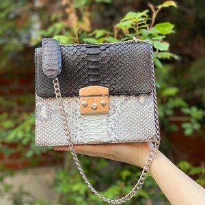 May include: A two-tone handbag with a snakeskin pattern. The top half is black, and the bottom half is white and gray. It has a silver chain strap and a gold-colored clasp.