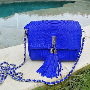 May include: A vibrant blue crossbody bag with a textured snakeskin pattern. The bag features a silver chain strap and a matching blue tassel. The text "AditReple" is visible on the front of the bag.