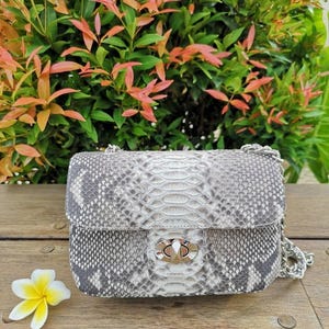 Puede incluir: Un bolso rectangular con estampado de piel de serpiente gris y blanco, con una correa de cadena plateada y un cierre. El bolso está sobre una superficie de madera con una flor blanca y amarilla en primer plano y follaje verde en el fondo.